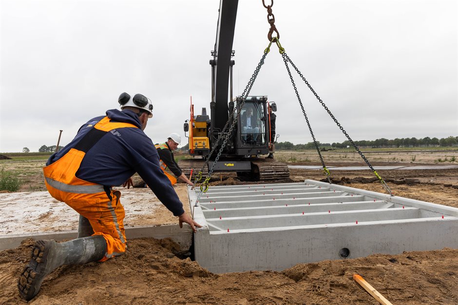 Twee mannen begeleiden een betonnen bak die met een kraan de grond in zakt, op de Hullenzandweg.