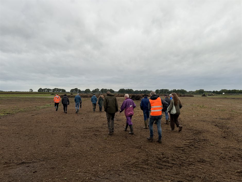 Een groep mensen loopt tijdens een excursie in Mantingerzand over kale grond. Verderop liggen stapels boomstronken.