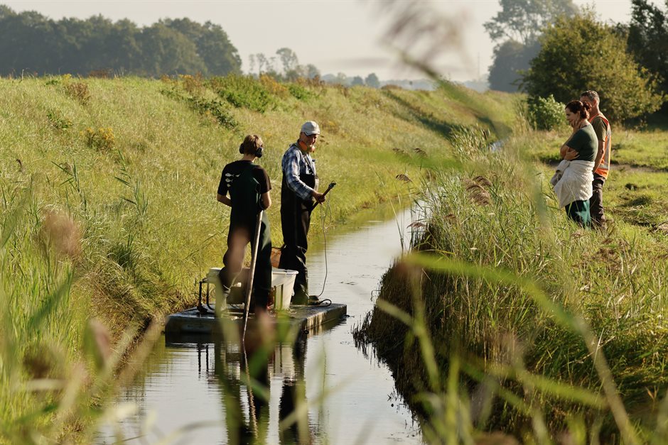 Twee mensen op een vlotje vangen vissen in een sloot in Laage Veen. Twee anderen kijken vanaf de kant toe.